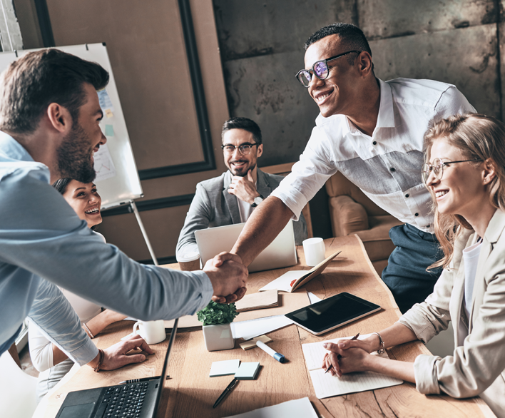 Group of individuals, three men and 2 women, in a meeting. Two of the men are shaking hands.