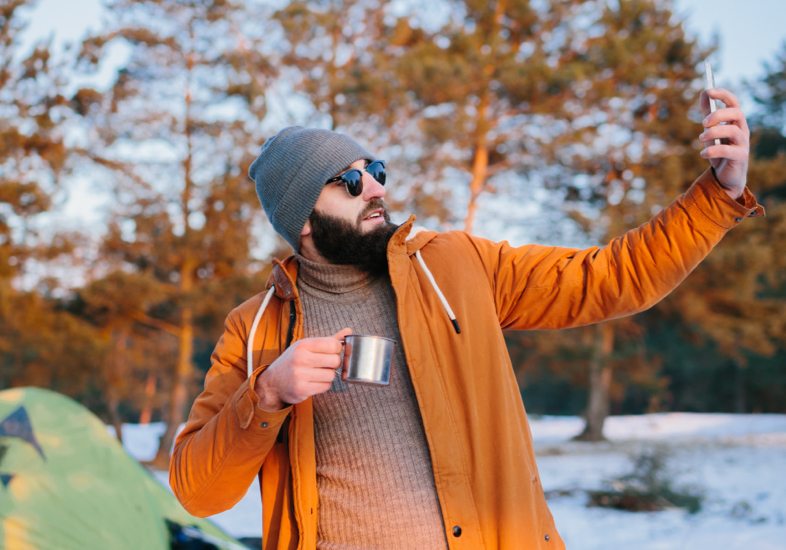 man on camping trip sitting in front of fire with cell phone on hand